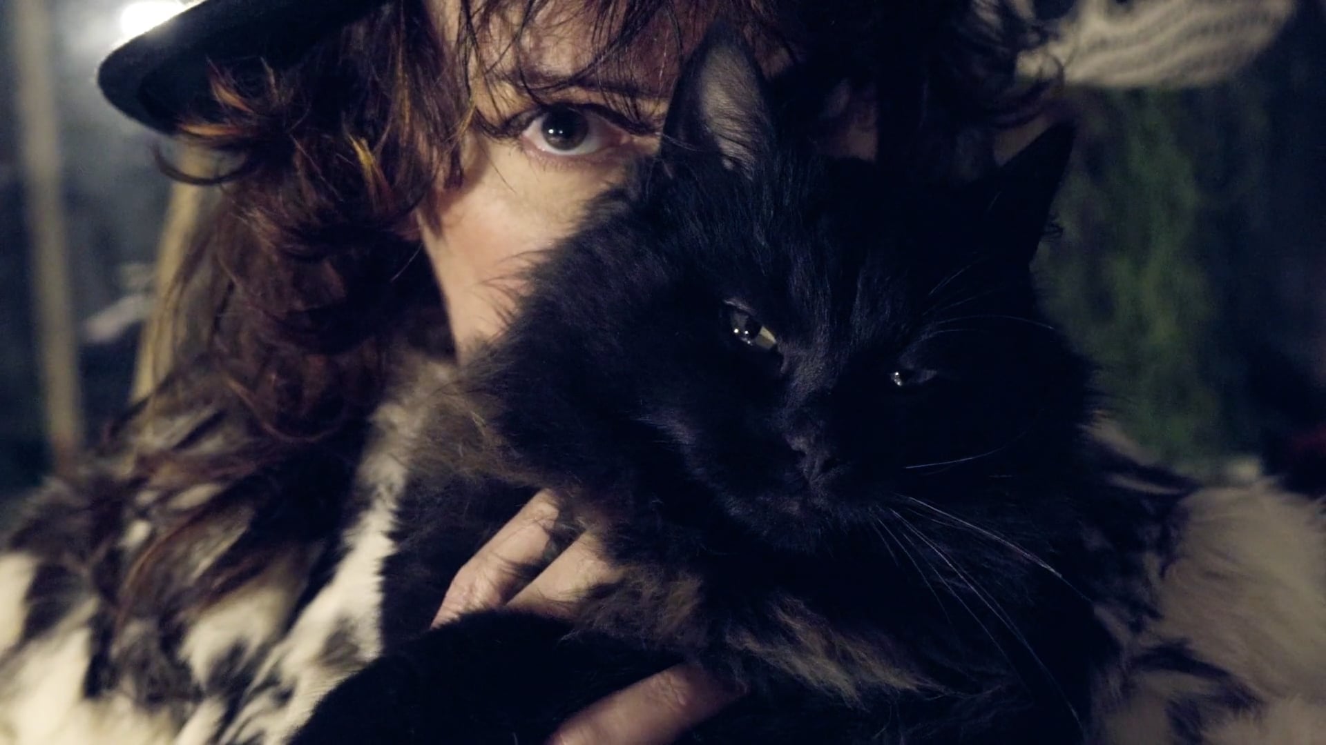 A person with tousled hair holds a black cat close to their face in dim, moody lighting