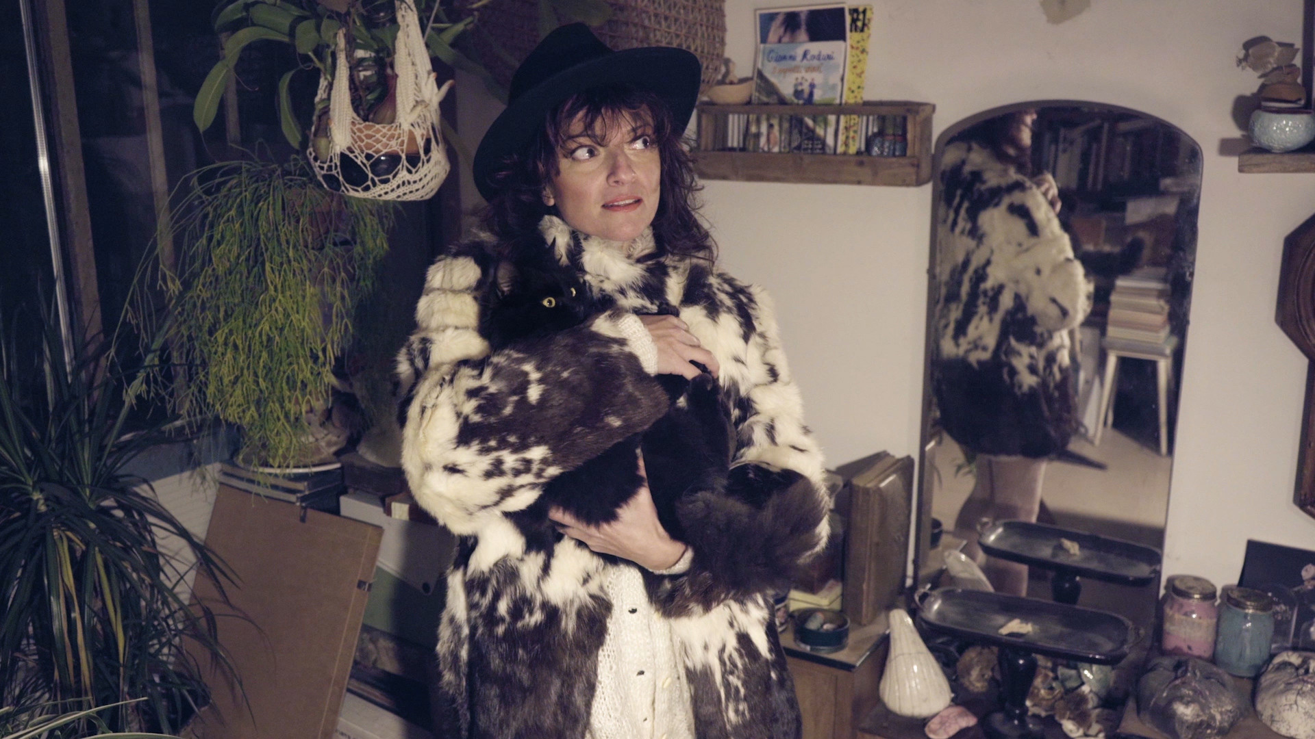 A woman in a fur coat and black hat holds a black cat against her chest, standing in a room with plants and a mirror reflecting her from behind